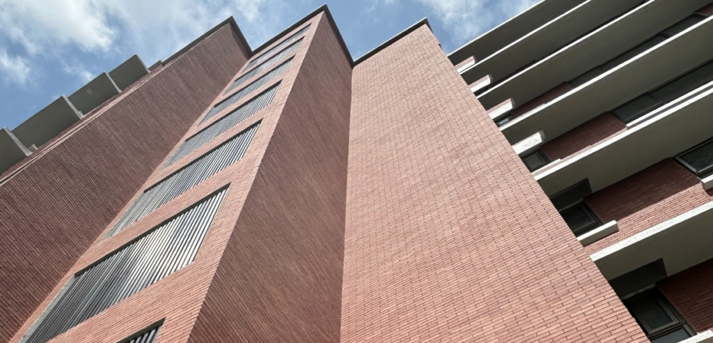 Low-angle view of a modern multi-storey building with red brick façade, vertical metal window louvers, and stacked balconies against a blue sky with light clouds — External Cladding Phomi MCM India.