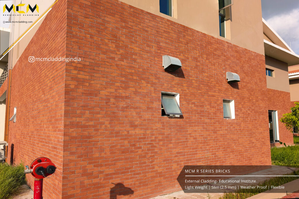A modern building’s exterior is clad in slim reddish-brown bricks. Three rectangular windows and four small ventilation boxes are set in the wall. The wall is sunlit, with a fire hydrant in the foreground and clear blue sky above. Text about the cladding appears.