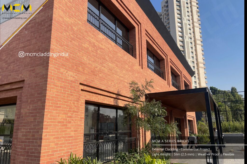 A modern commercial building features red brick cladding, large windows with black frames, and a black metal awning. Green shrubs and trees line the base. A tall high-rise building is visible in the background under a clear blue sky.