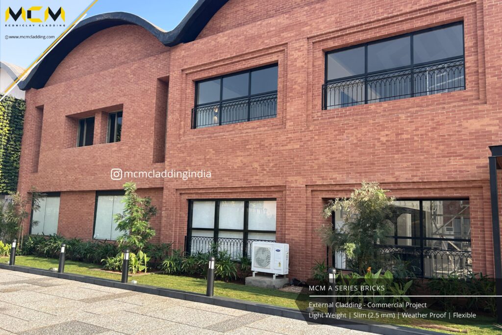 A modern two-story building features reddish-brown brick cladding, large rectangular windows with black frames, and a wavy roofline. Green shrubs and small trees line the front, with a paved walkway and a white outdoor air conditioning unit visible.