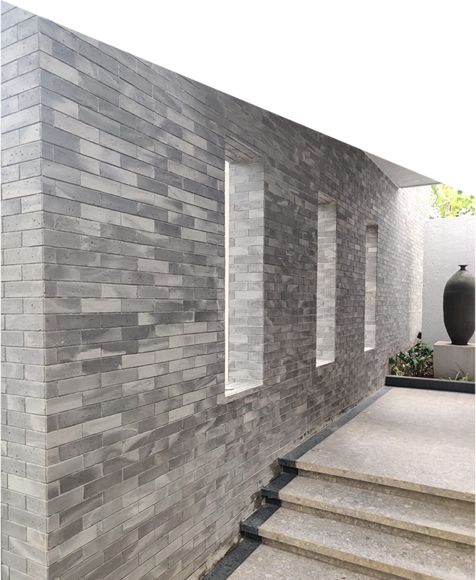 A modern exterior wall made of gray bricks features three narrow vertical windows. Stone steps lead upward along the wall, and a large black urn decorates a minimalist courtyard with some greenery in the background. The setting is bright and clean.