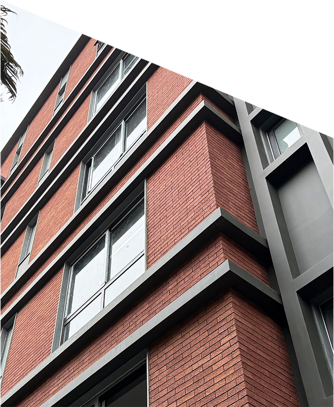 A modern multi-story building with red brick walls and gray metal framing, viewed from below. Large rectangular windows are evenly spaced across each floor, and some palm tree leaves are visible in the upper left corner against a pale sky.